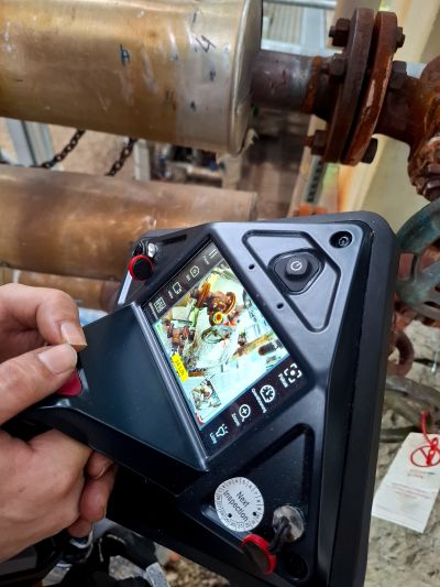 Hand hält eine Ultraschallkamera vor einer rostigen Flanschverbindung, Display zeigt die akustische Overlay-Darstellung